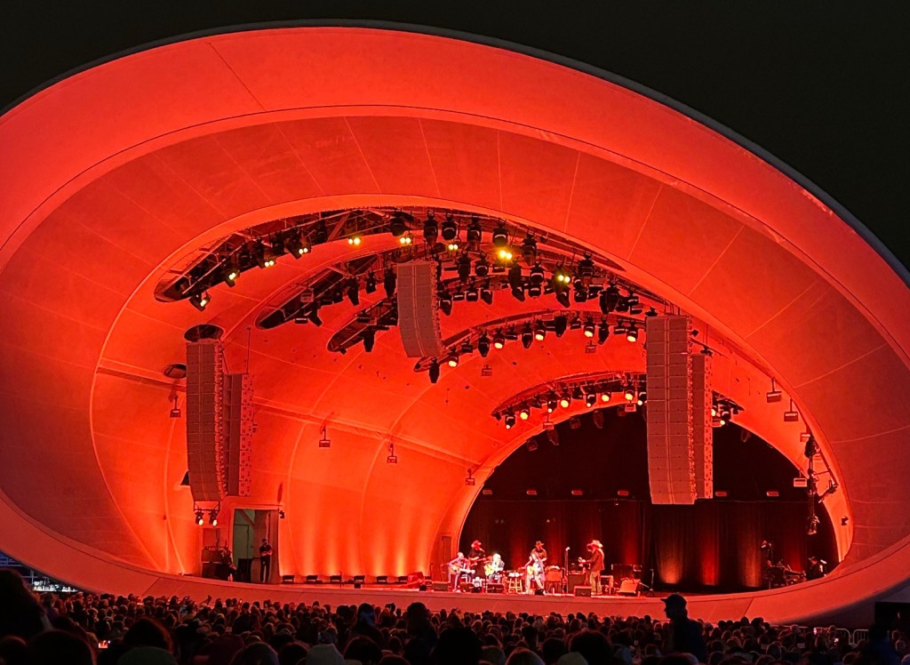 outdoor concert venues in San Diego; Rady Shell is a big conical shaped structure making up an outdoor venue. It glows red. Willie Nelson is on stage playing.
