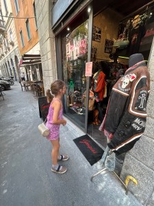 A young girl in a purple outfit looks at a vintage clothing store display featuring a jacket in Milan.