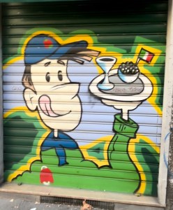 Colorful graffiti mural of a smiling waiter holding a tray with a drink and food, depicted on a green garage door.